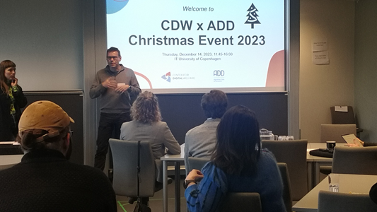 2 presenters standing in front of an audience in front of an image saying "welcome to CDW x ADD Christmas Event 2023"